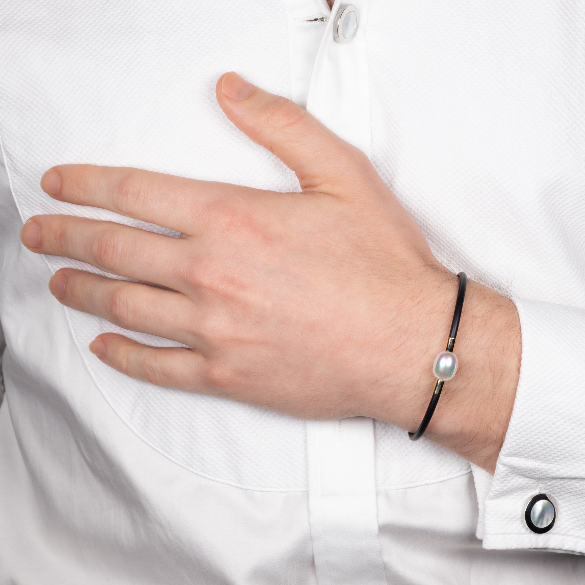 Hand wearing a black bracelet with a pearl on a white shirt background