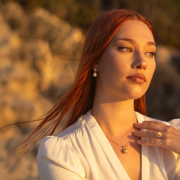 Woman with red hair wearing a white top and gold pearl jewelry against a blurred natural background
