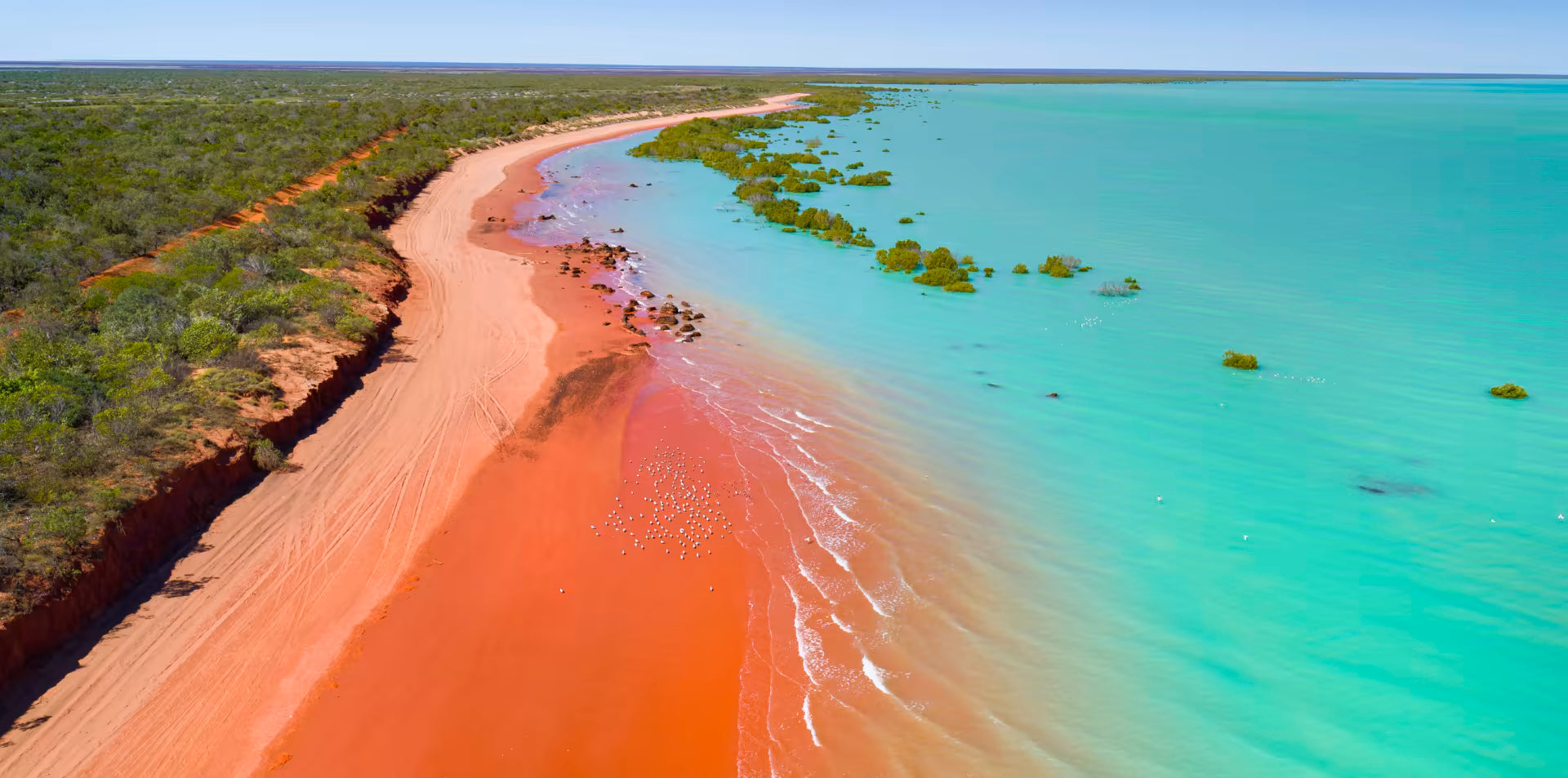 Aerial view of a coastal landscape with red sand, clear blue water, and greenery.