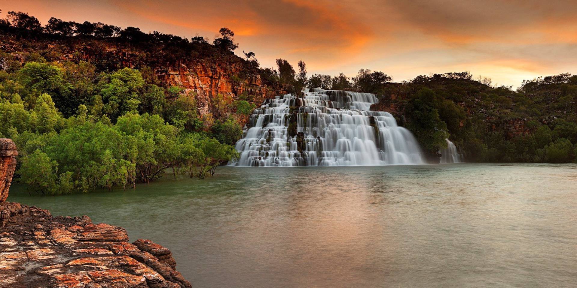 Waterfall cascading into a lake with a sunset sky and rocky landscape.